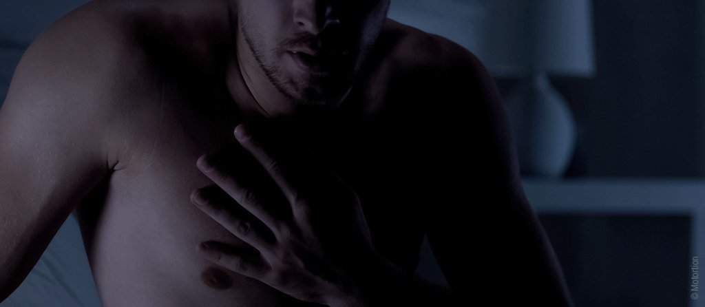 Photo : un homme torse nue est assis dans une pièce sombre et se touche la poitrine.