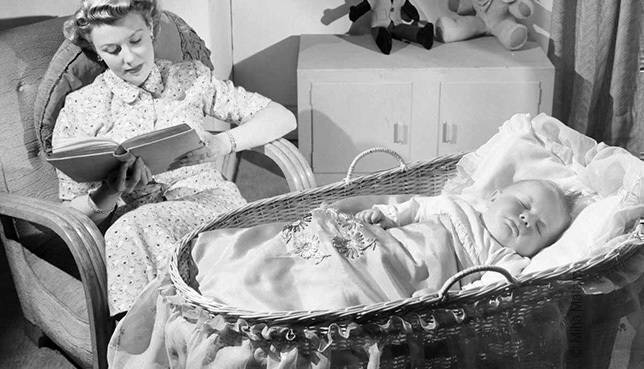 Photo : un bébé est couché dans un berceau. En arrière-plan, la mère est assise et lit un livre pour bien dormir