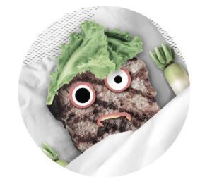 Une escalope avec des yeux en rondelles de radis, des bras en poireaux et des cheveux en laitue iceberg est à moitié couverte dans un lit.