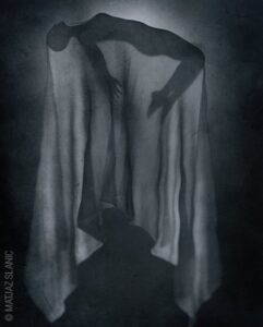 Photo : une silhouette en lévitation est recouverte d'un drap dans une pièce noire. ses bras et ses jambes pendent.