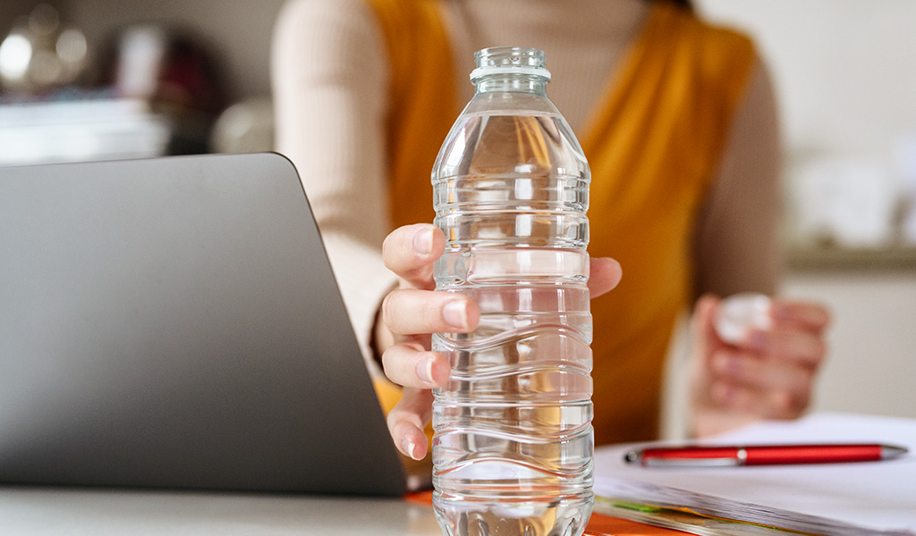 Une bouteille d'eau en gros plan est tenue par une main de femme floutée dans le fond.