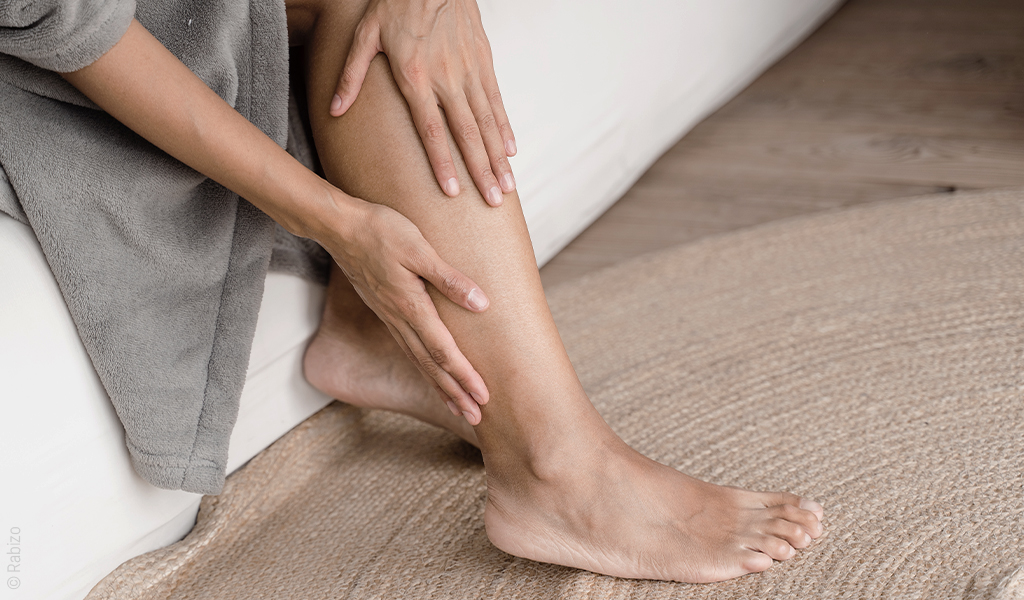 des mains féminines se tiennent les jambes. Les pieds sont sur un tapis en jute.