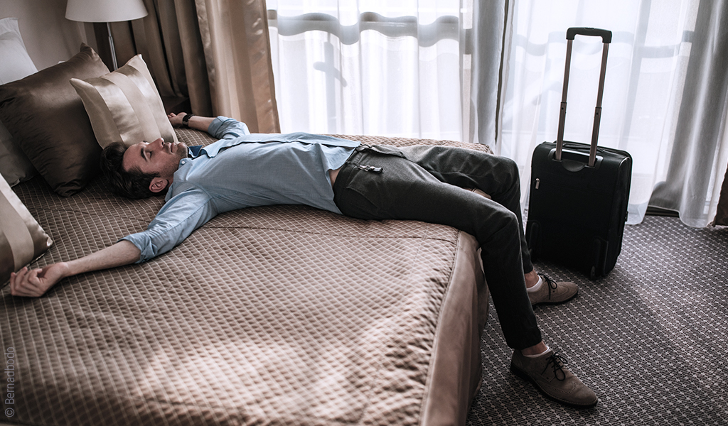 Un homme est avachi à moitié sur son lit dans une chambre d'hôtel. Une valise est près de son lit.